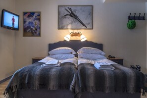 Un letto matrimoniale con testiera, una televisione a sinistra, un quadro incorniciato sopra il letto, una lampada a parete e un appendiabiti con un casco.