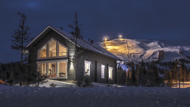 Uma casa moderna de dois andares, com janelas amplas e entrada bem iluminada, cercada por neve e árvores, com uma montanha ao fundo.