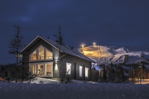A modern two-story house with large windows and a well-lit entrance, surrounded by snow and trees, with a mountain in the background. - Trillevallen Högfjällshotell (Undersaker)