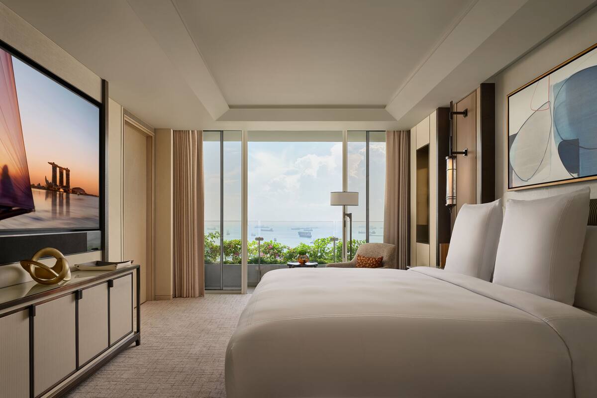 A hotel room with a large bed, a TV, a view of the sea, and a balcony with curtains.