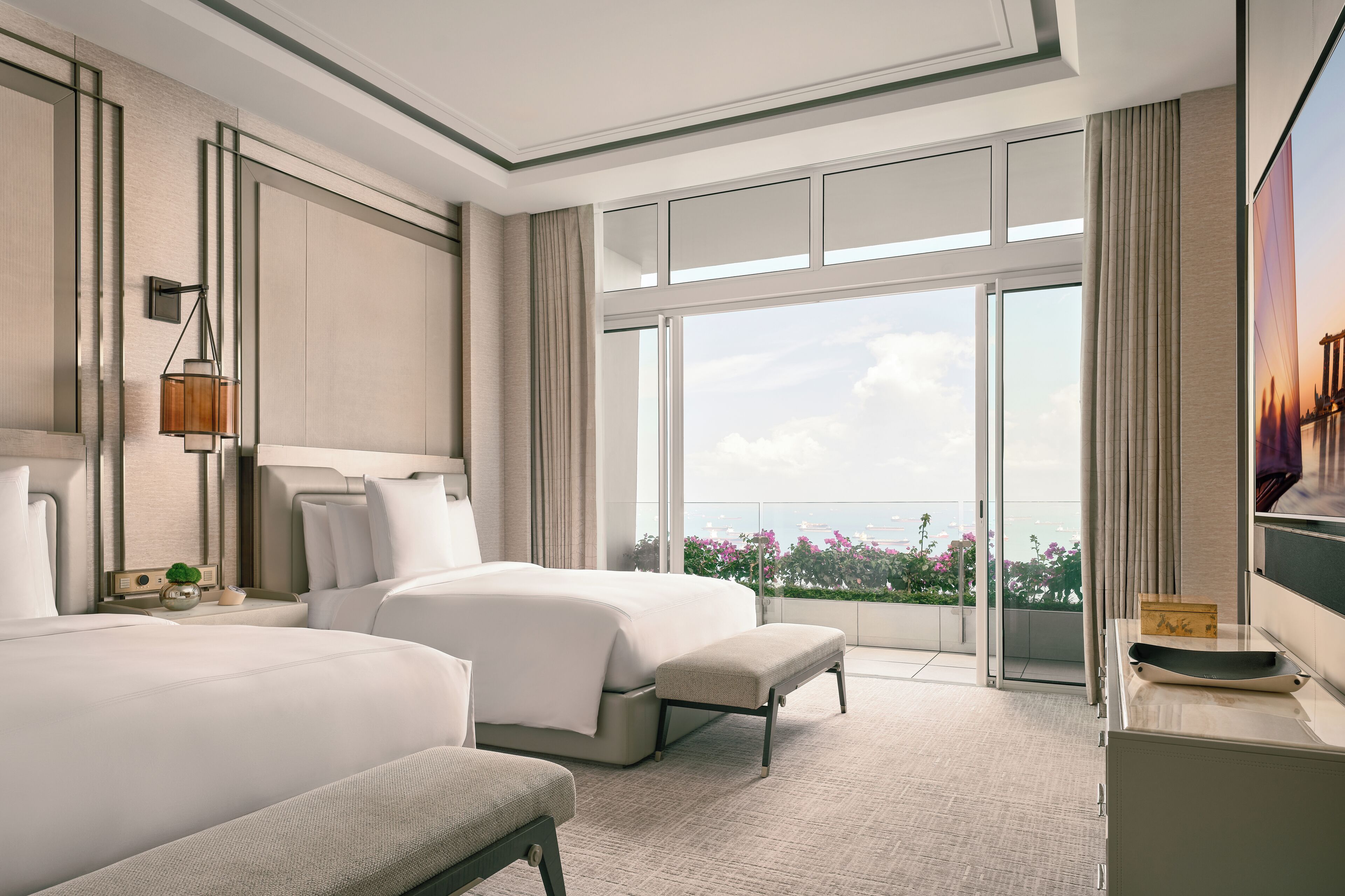 A modern hotel room with two beds, a large window with a city view, a flat-screen TV, and a balcony with flowers.