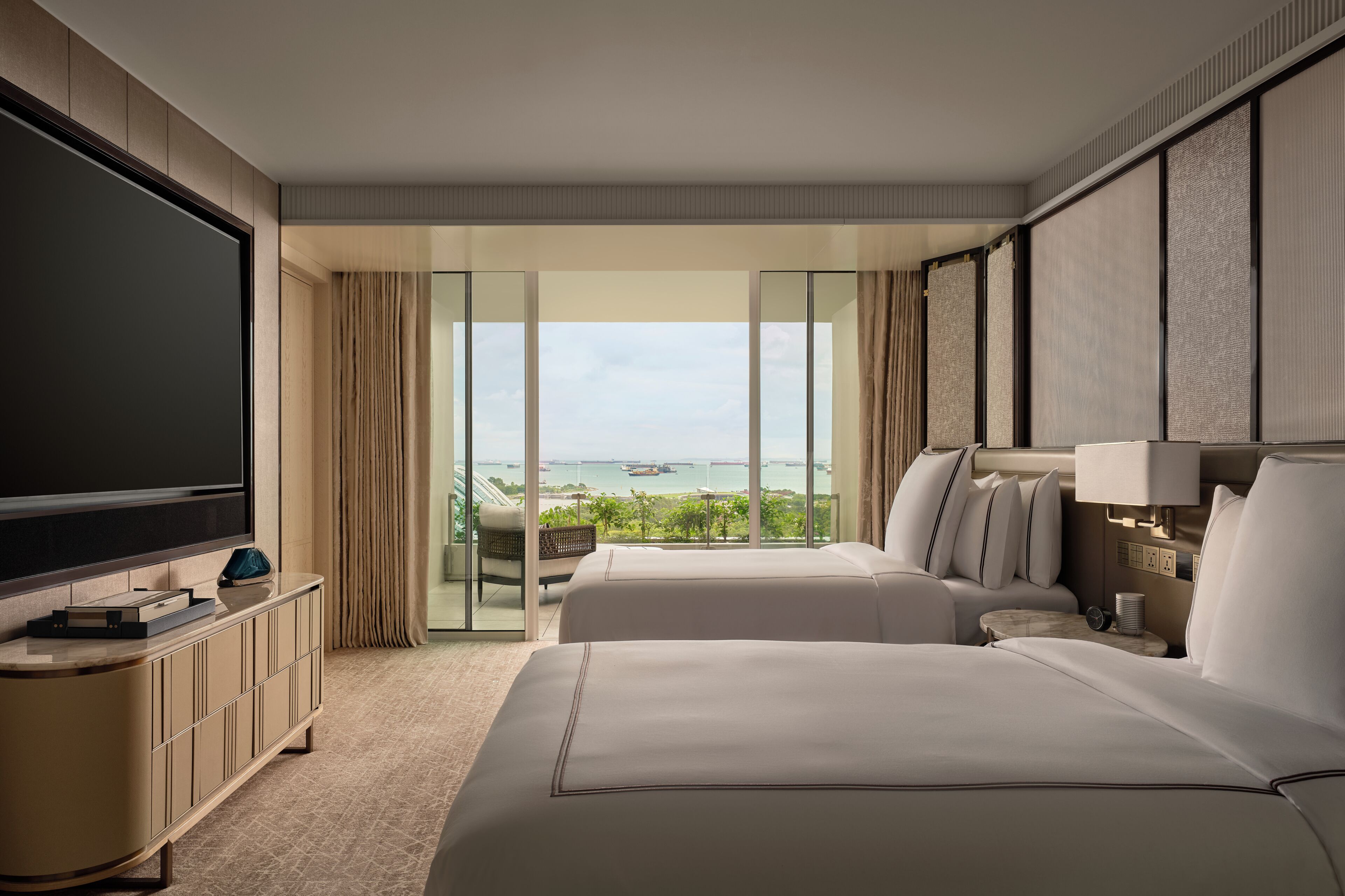 A hotel room with two beds, a TV, a large window with a view of the sea, and a balcony with curtains.