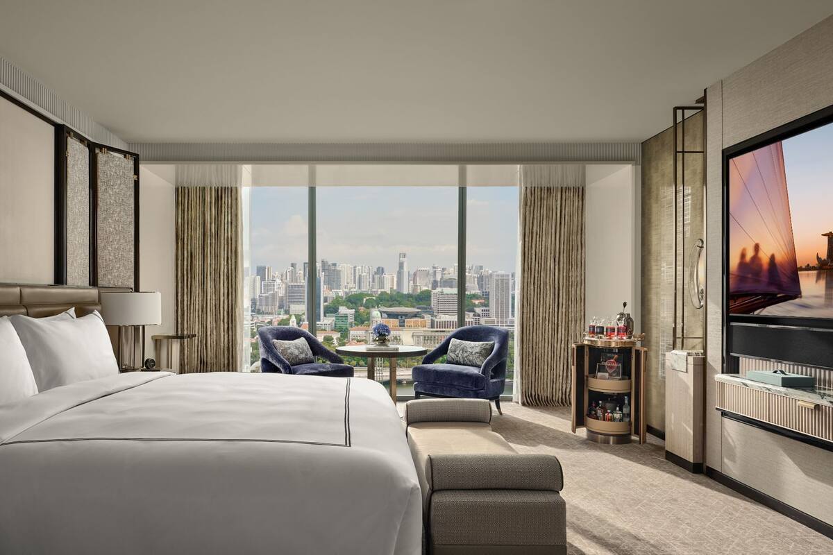 A hotel room with a large bed, two armchairs, a small table, a TV, and a city view.