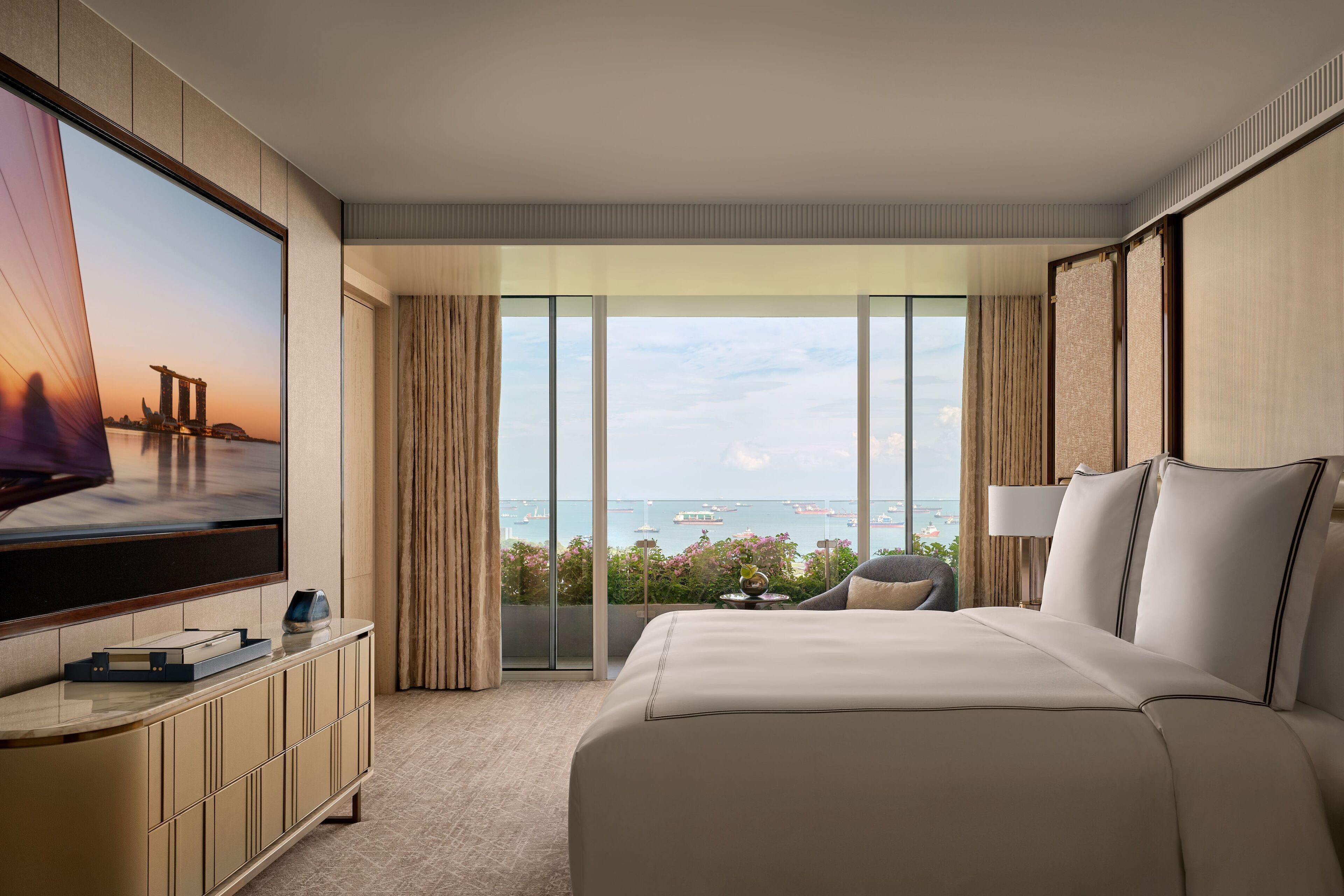 A hotel room with a large bed, a TV on a cabinet, and a balcony with a view of the sea.
