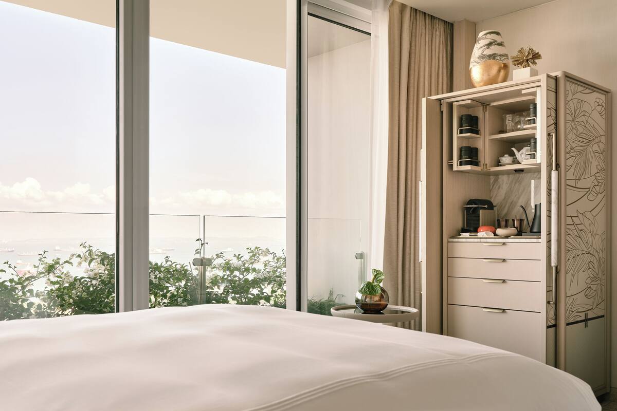 A hotel room with a large bed, a small table with a vase, a cabinet with drawers, and a view of greenery and a cityscape.