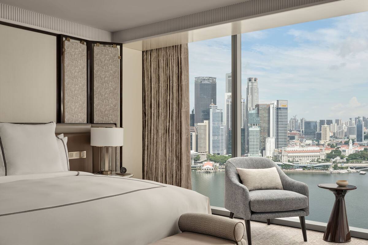 A modern hotel room with a large bed, a seating area with a chair and a small table, and a view of a city skyline and water.
