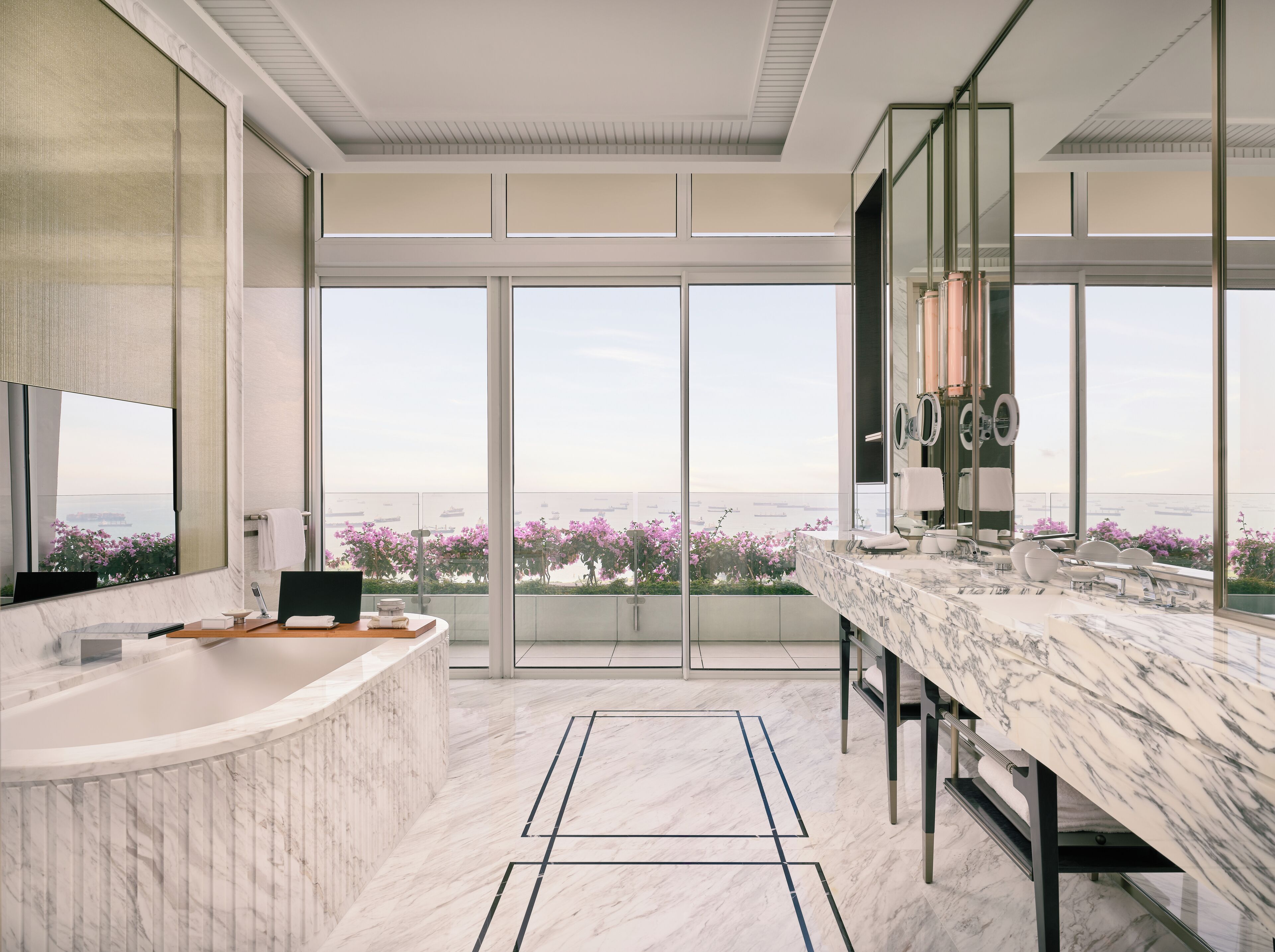 A modern bathroom with a large bathtub, a marble countertop, and a view of pink flowers and a distant ship.