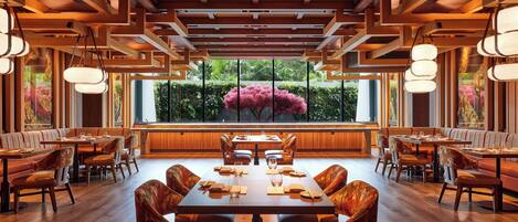 A restaurant with wooden ceiling beams, large windows with a view of greenery, and a central dining table with chairs.