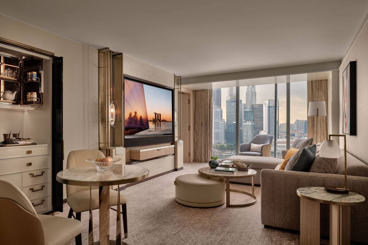 A modern hotel room with a large TV, comfortable seating, a dining area, and a city view.