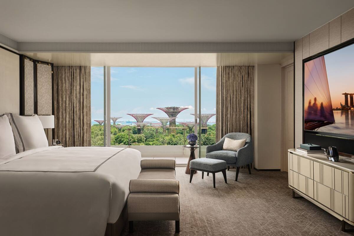 A hotel room with a large bed, a seating area, a television, and a view of a cityscape with distinctive architecture.