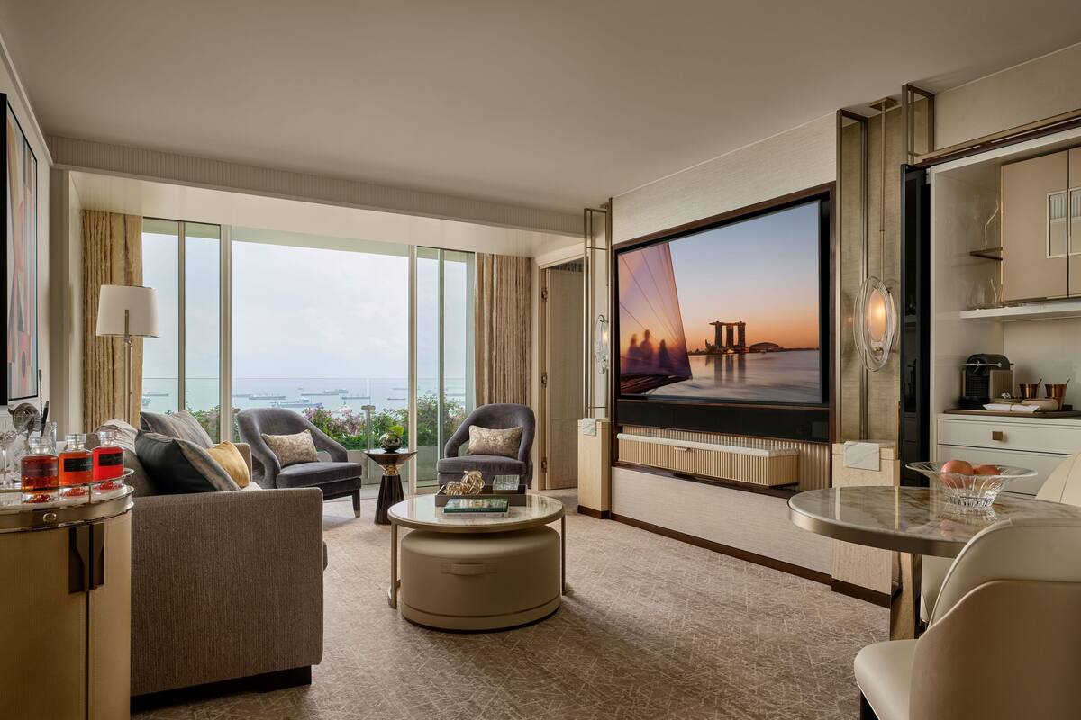 A spacious hotel room with a large TV, comfortable seating, a dining area, and a view of the cityscape.