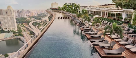 Una piscina con una persona en el agua, rodeada de sillones de playa y palmeras, con un paisaje urbano al fondo.