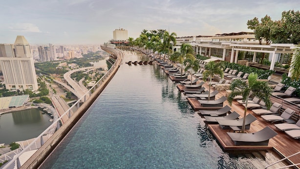 Una piscina con una persona en el agua, rodeada de sillones de playa y palmeras, con un paisaje urbano al fondo.