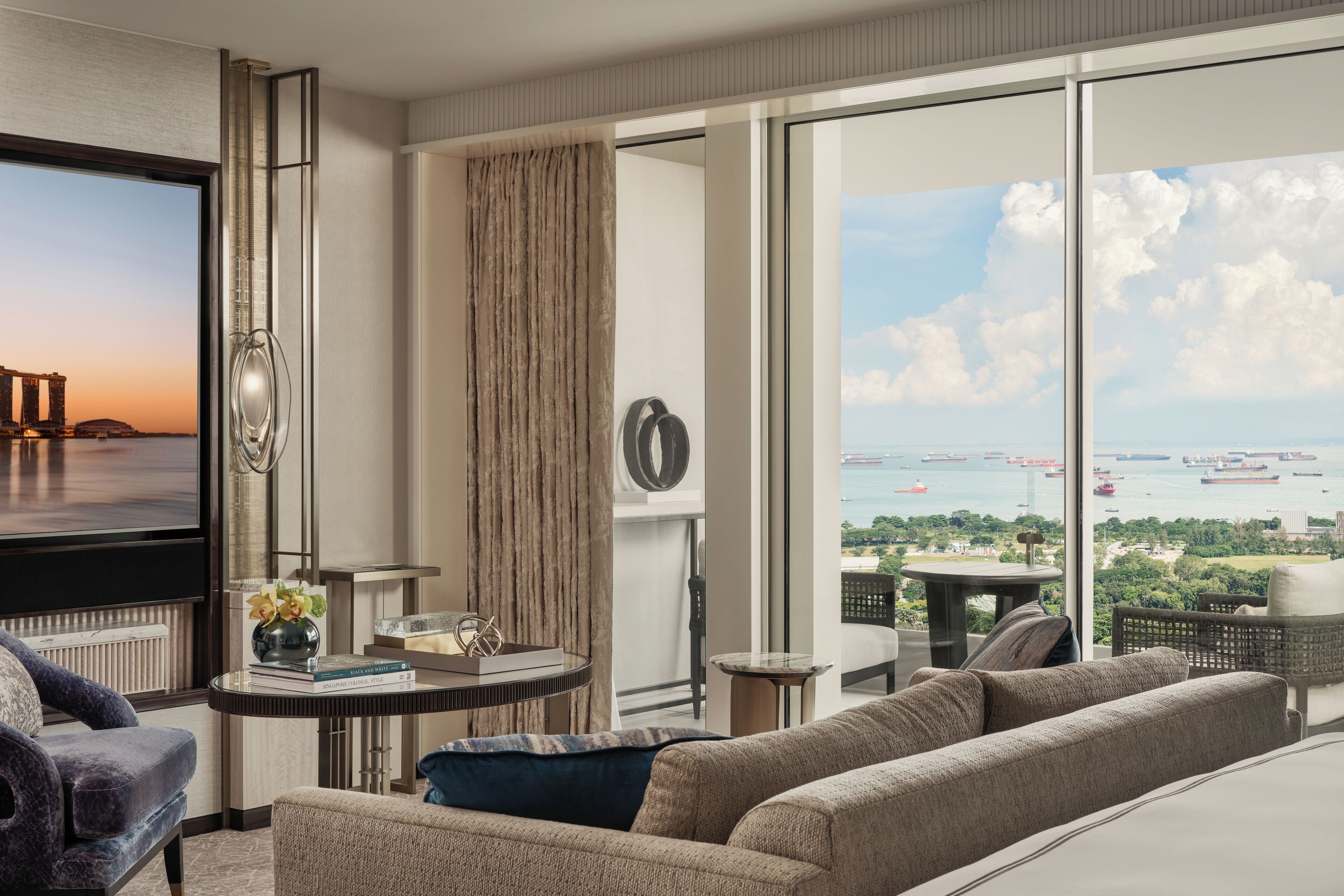 Habitación de hotel moderna con un televisor grande, asientos cómodos, una mesita con libros y vistas al mar y barcos.