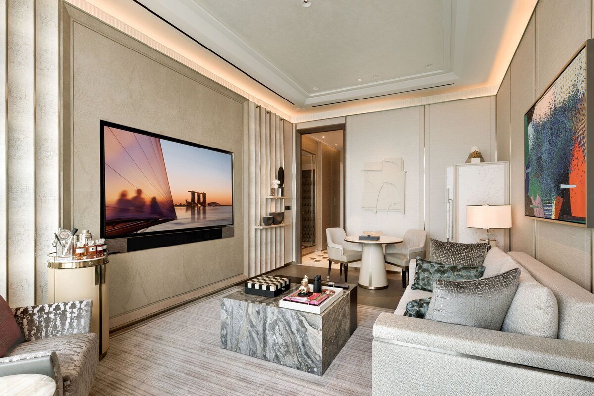 A modern hotel room with a large flat-screen TV, a marble coffee table, a sofa, a small round table, and a painting on the wall.