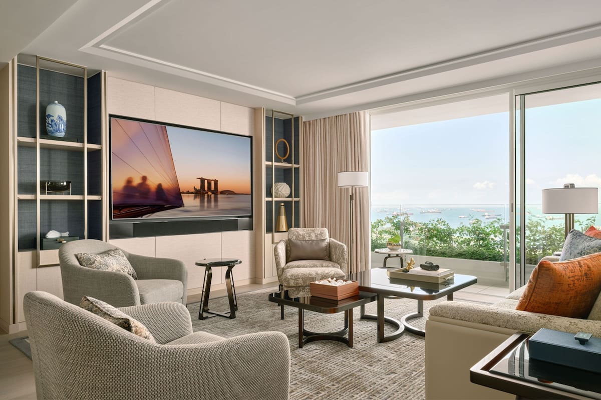 A modern hotel room with a large TV, comfortable seating, a view of the sea, and a balcony with a table and chairs.