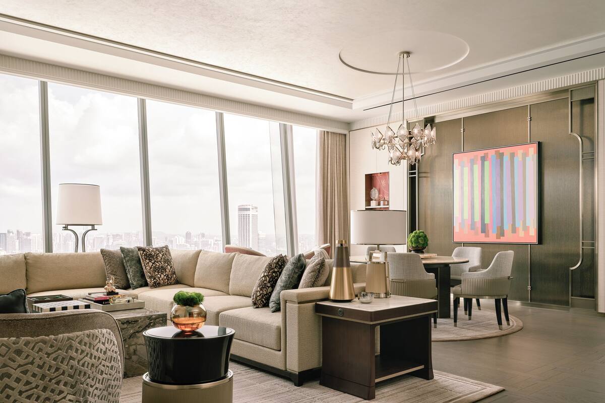 A modern living room with a sofa, coffee table, and chairs. There is a chandelier, a painting, and a city view through large windows.