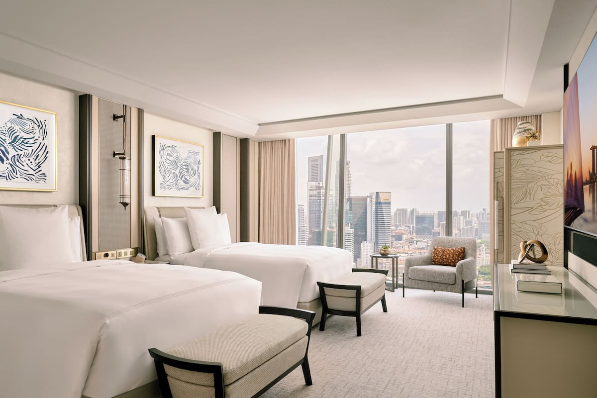 A hotel room with two beds, a desk, a chair, and a view of a cityscape.