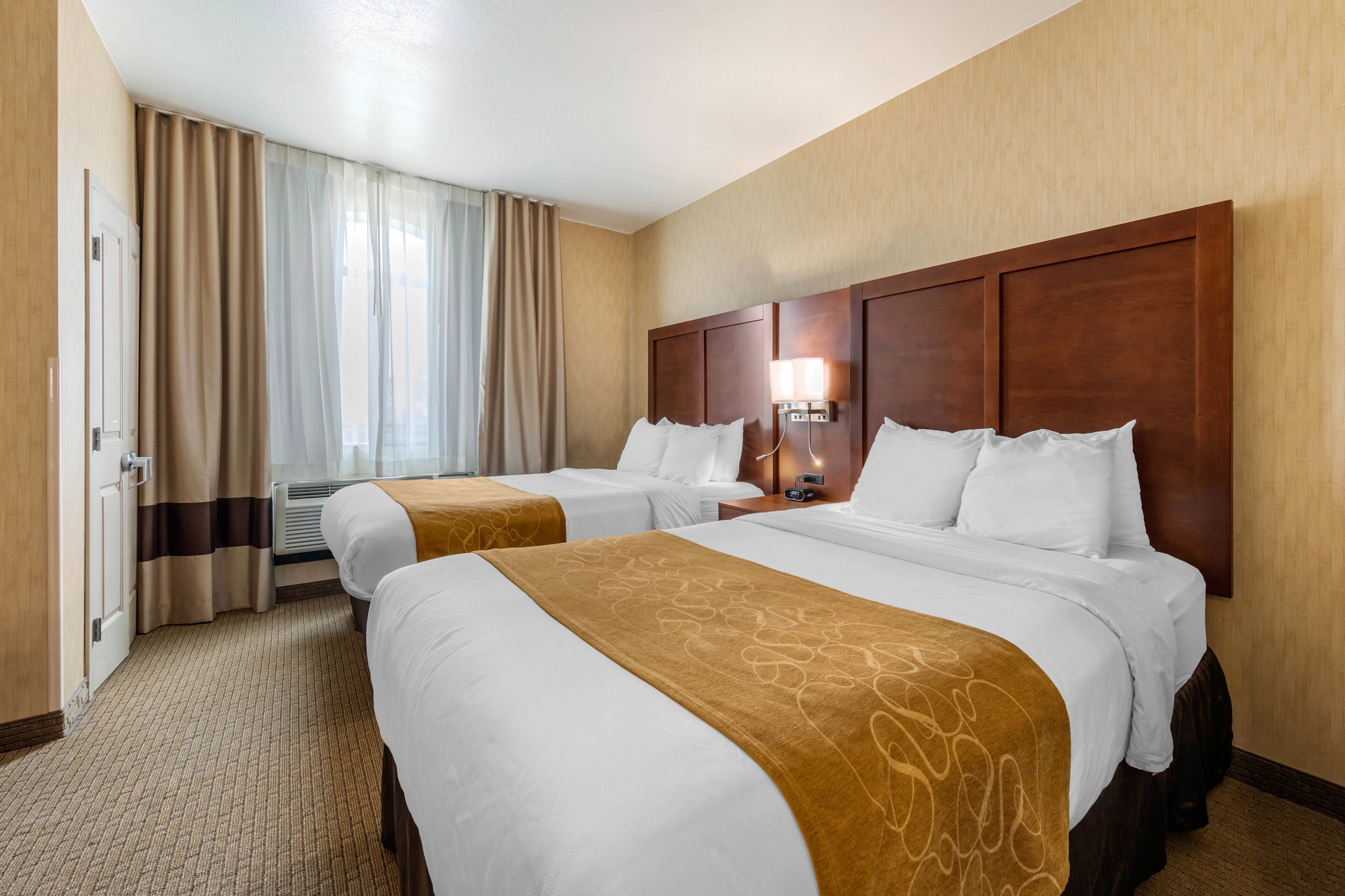 A hotel room with two beds, a wooden headboard, a bedside table with a lamp, and a window with curtains.