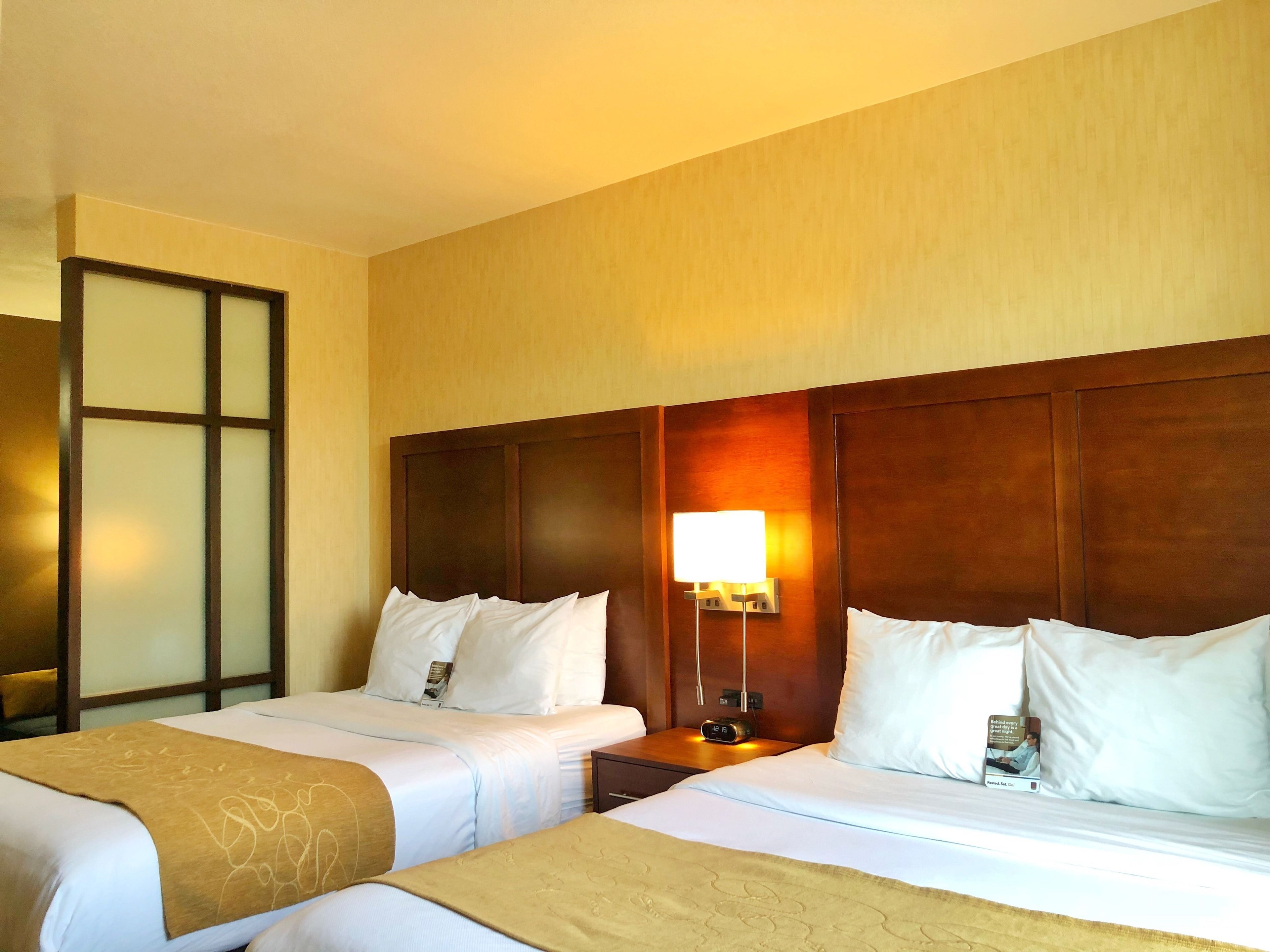 A hotel room with two beds, a nightstand, a lamp, and a mirror.