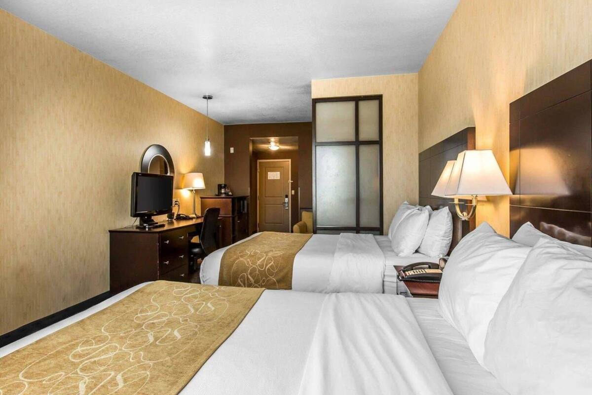 A hotel room with two beds, a desk with a computer, a chair, a lamp, and a door leading to another room.