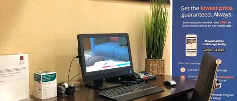 Balcão de recepção de hotel com computador, fotografia emoldurada de um panorama urbano, planta e um cartaz promocional.