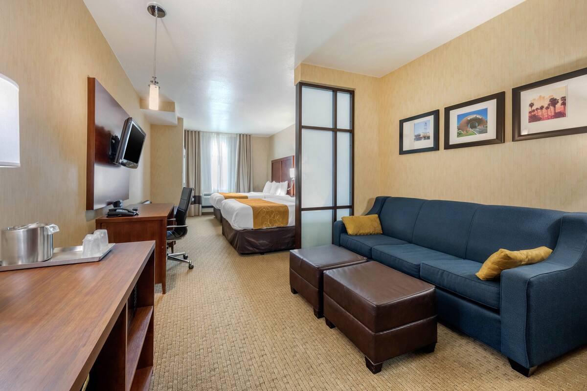 A hotel room with a wooden desk, a blue sofa, a bed with a white and brown bedspread, a television mounted on the wall, and framed pictures on the wall.
