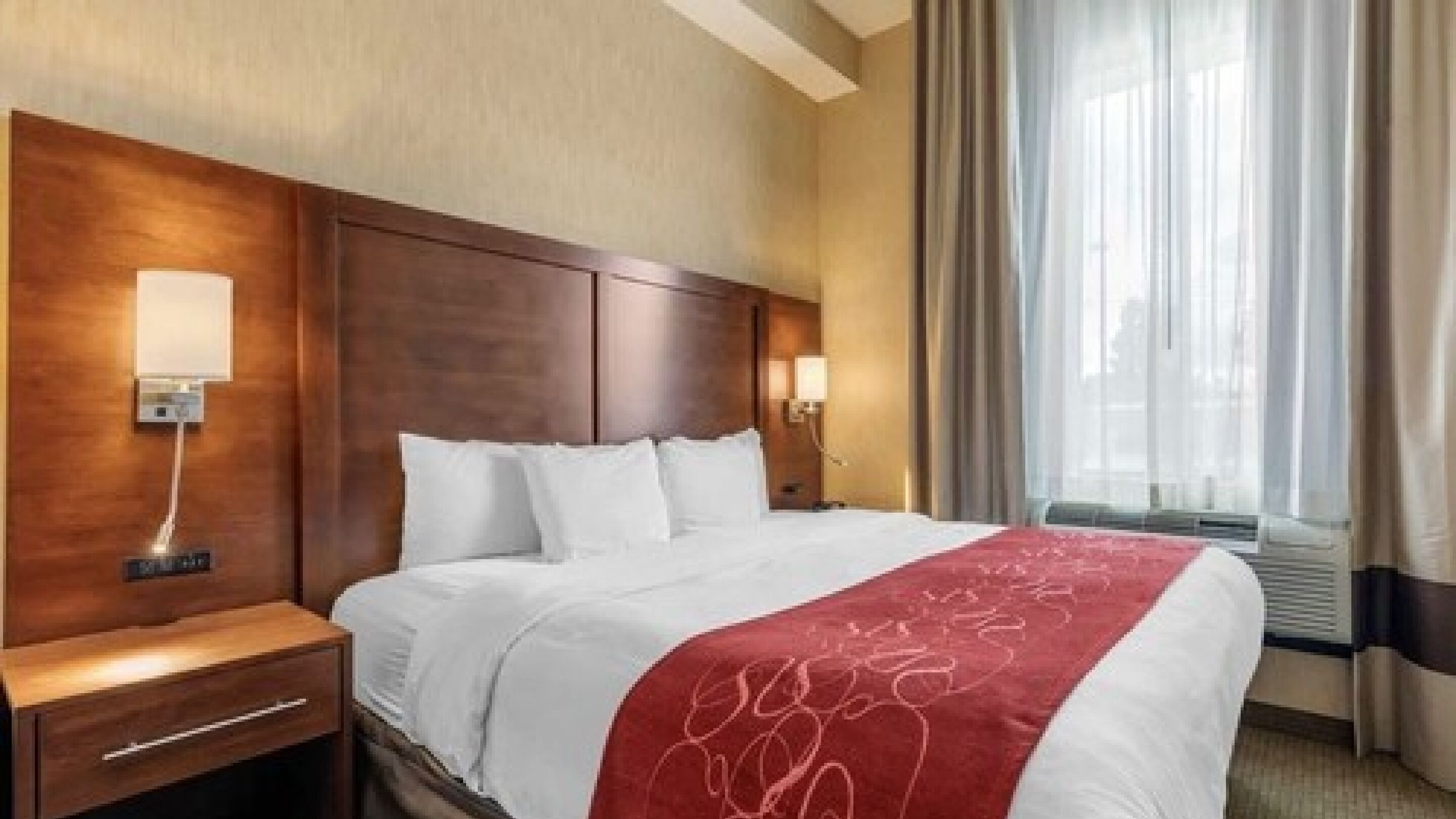 Quarto de hotel com uma cama grande, duas mesas de cabeceira, um painel de cama de madeira e uma janela com cortinas.