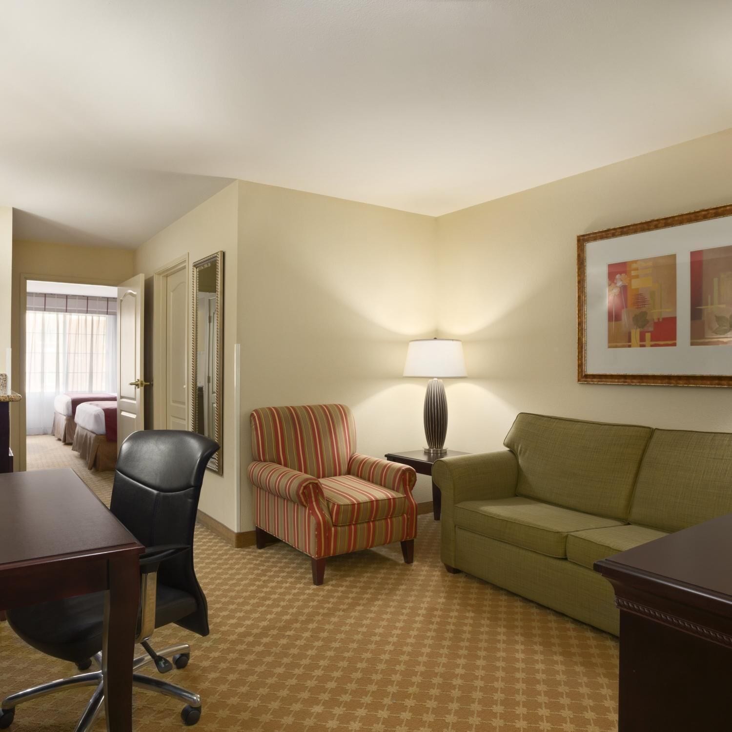 A hotel room with a desk, a striped armchair, a green sofa, a lamp, and a framed picture on the wall.