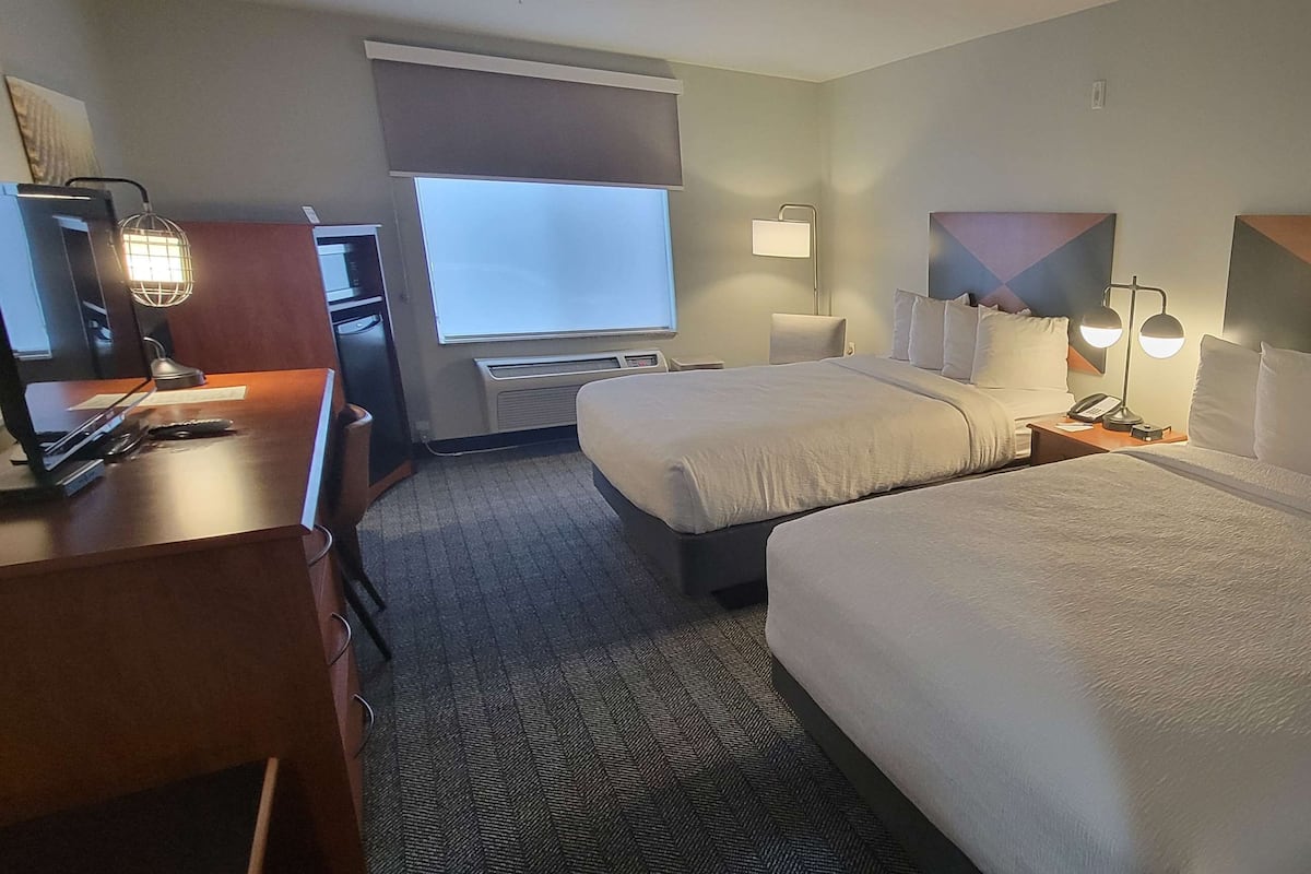 A hotel room with two beds, a desk, a chair, a lamp, and a window.