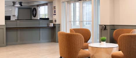 A lobby with a reception desk, two orange chairs around a small table, and a large window with a view.