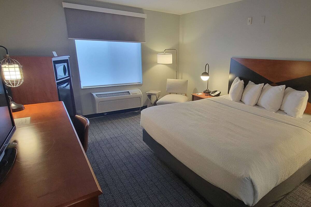 A hotel room with a large bed, a desk, a chair, a desk lamp, a wall-mounted TV, and a window with blinds.