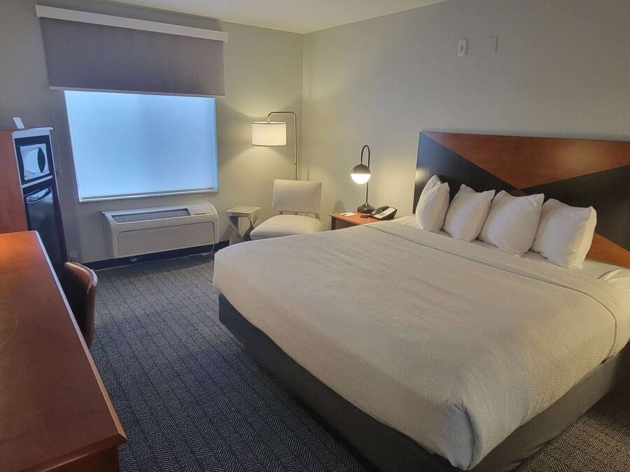 Habitación de hotel con una cama grande, un escritorio, una silla, una lámpara de escritorio, un televisor de pared y una ventana con persianas.