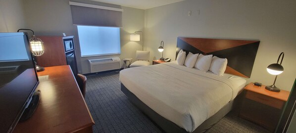 A hotel room with a large bed, a desk, a chair, a desk lamp, a wall-mounted TV, and a window with blinds.