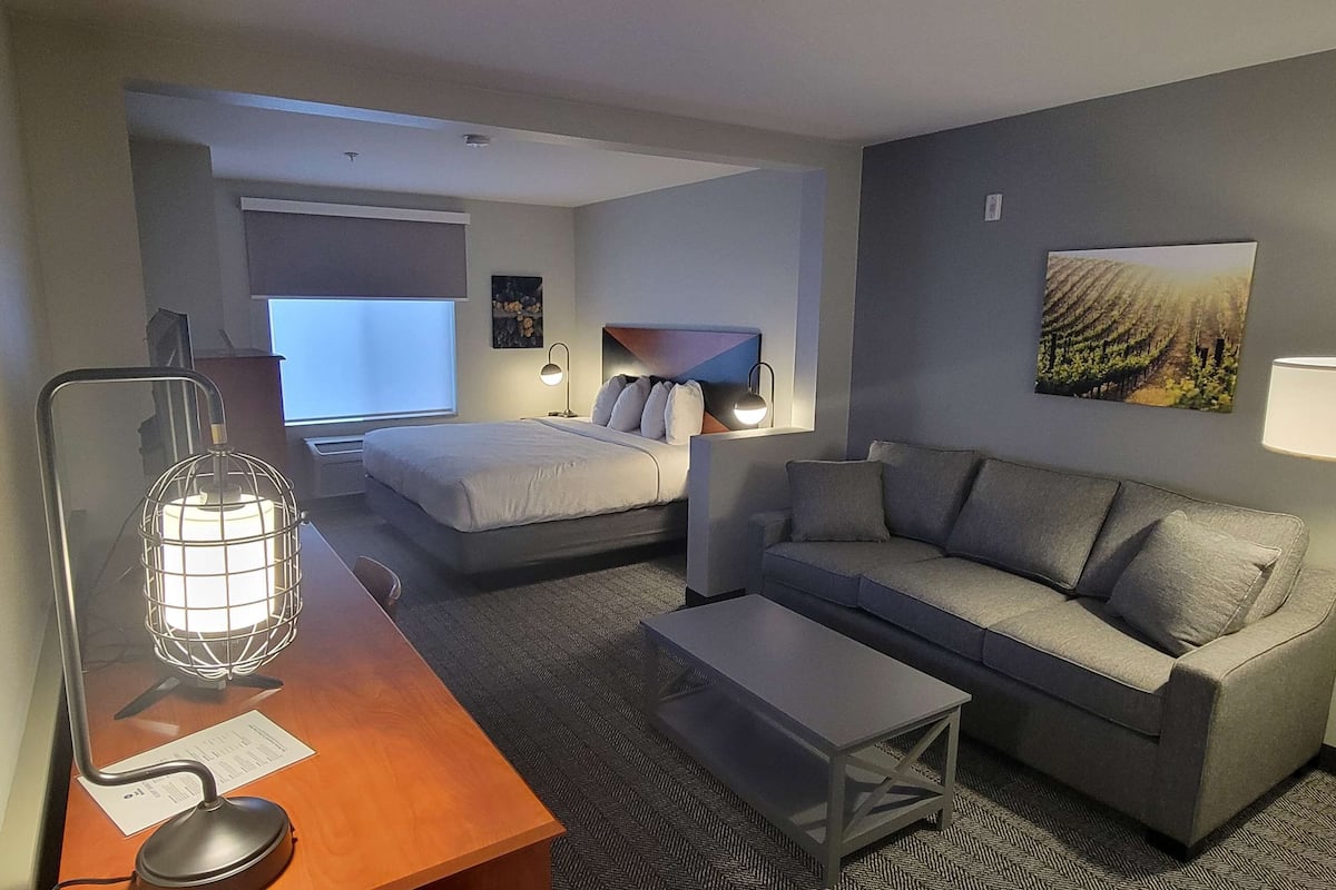 A hotel room with a bed, a sofa, a desk with a lamp, and a painting on the wall.