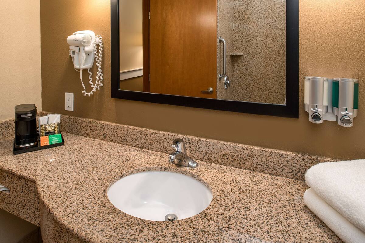 A hotel bathroom with a granite countertop sink, a mirror, a hairdryer, and toiletries.