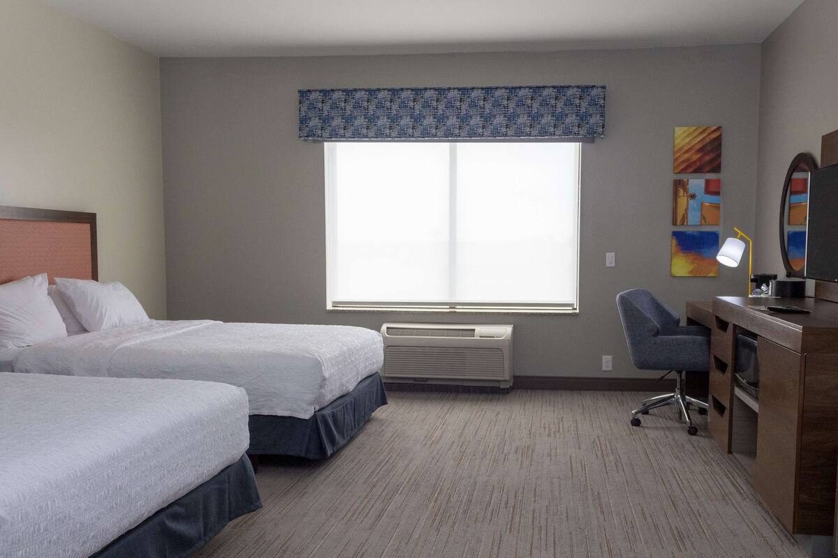 Ein Hotelzimmer mit zwei Betten, einem Schreibtisch mit Stuhl, einem großen Fenster mit Jalousien, einer Klimaanlage und Bildern an der Wand.