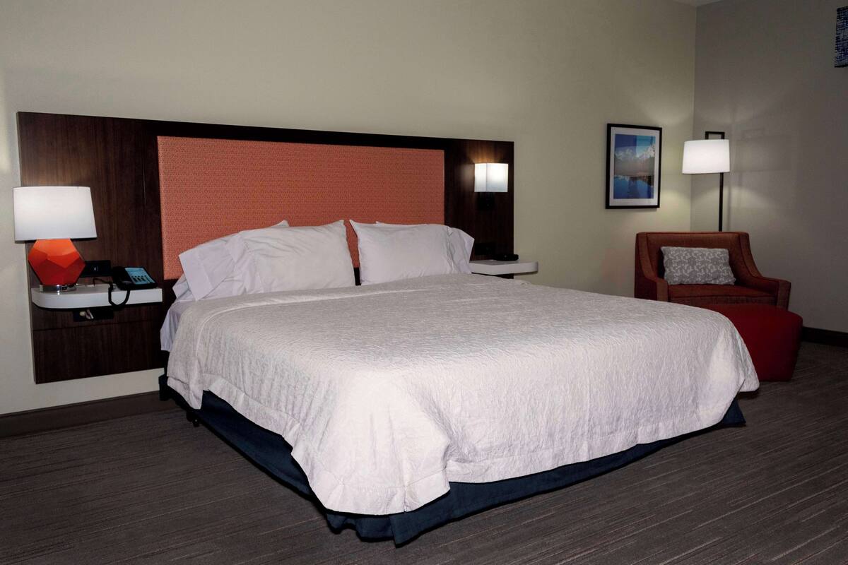 Ein Hotelzimmer mit einem großen Bett, einem Nachttisch mit Telefon und Lampe, einem Sessel mit Kissen und einem gerahmten Bild an der Wand.