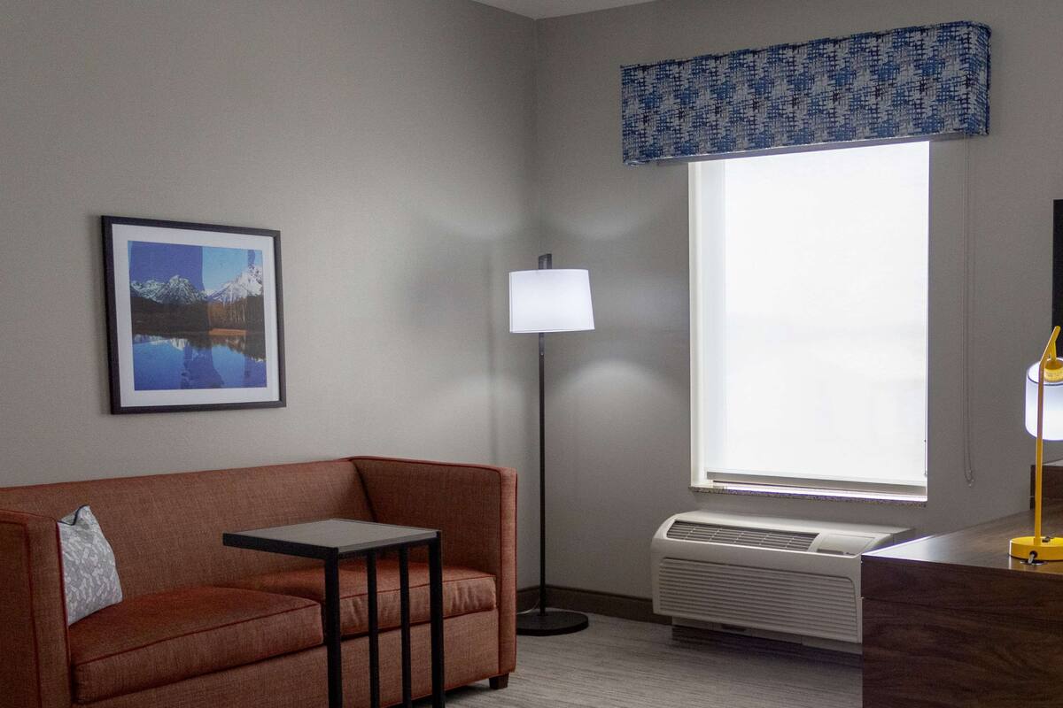 Ein Hotelzimmer mit Sofa, kleinem Tisch, Lampe, Fenster mit Blick und Heizung.