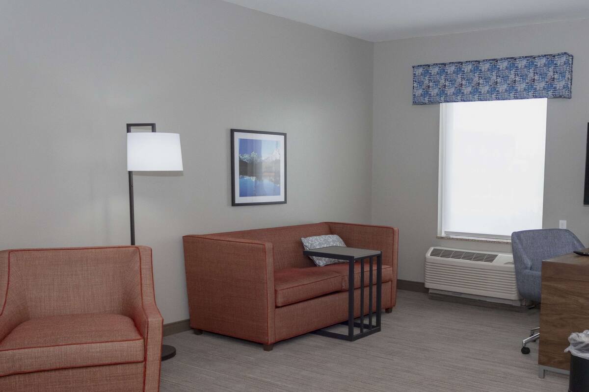 Ein modernes Wohnzimmer mit Sofa, Sessel und einem kleinen Tisch. An der Wand hängt ein gerahmtes Bild, eine Stehlampe steht bereit und ein Fenster ist mit einem blauen Vorhang verziert.