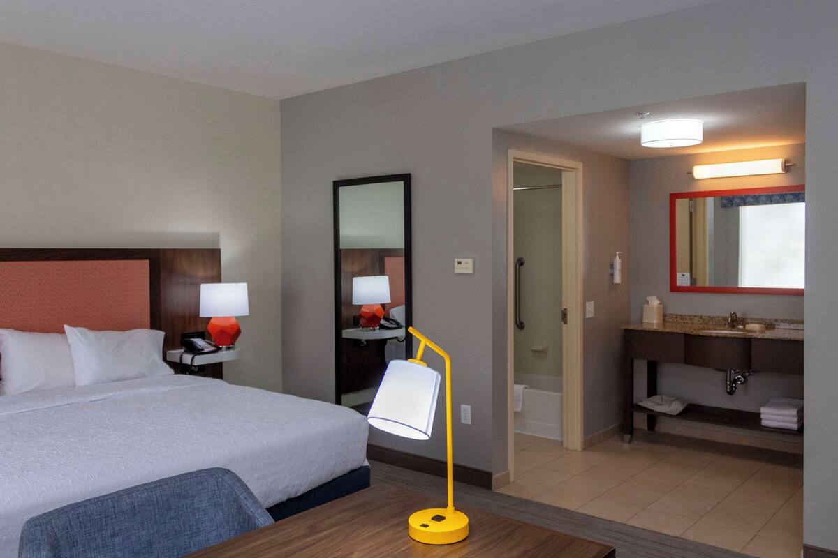 Ein Hotelzimmer mit Bett, Nachttisch mit Lampe, Spiegel, Badezimmer mit Waschbecken und Spiegel sowie einer Tür zu einem anderen Raum.