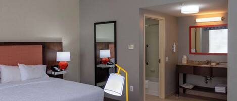 A hotel room with a bed, bedside table with a lamp, a mirror, a bathroom with a sink and mirror, and a door leading to another room.
