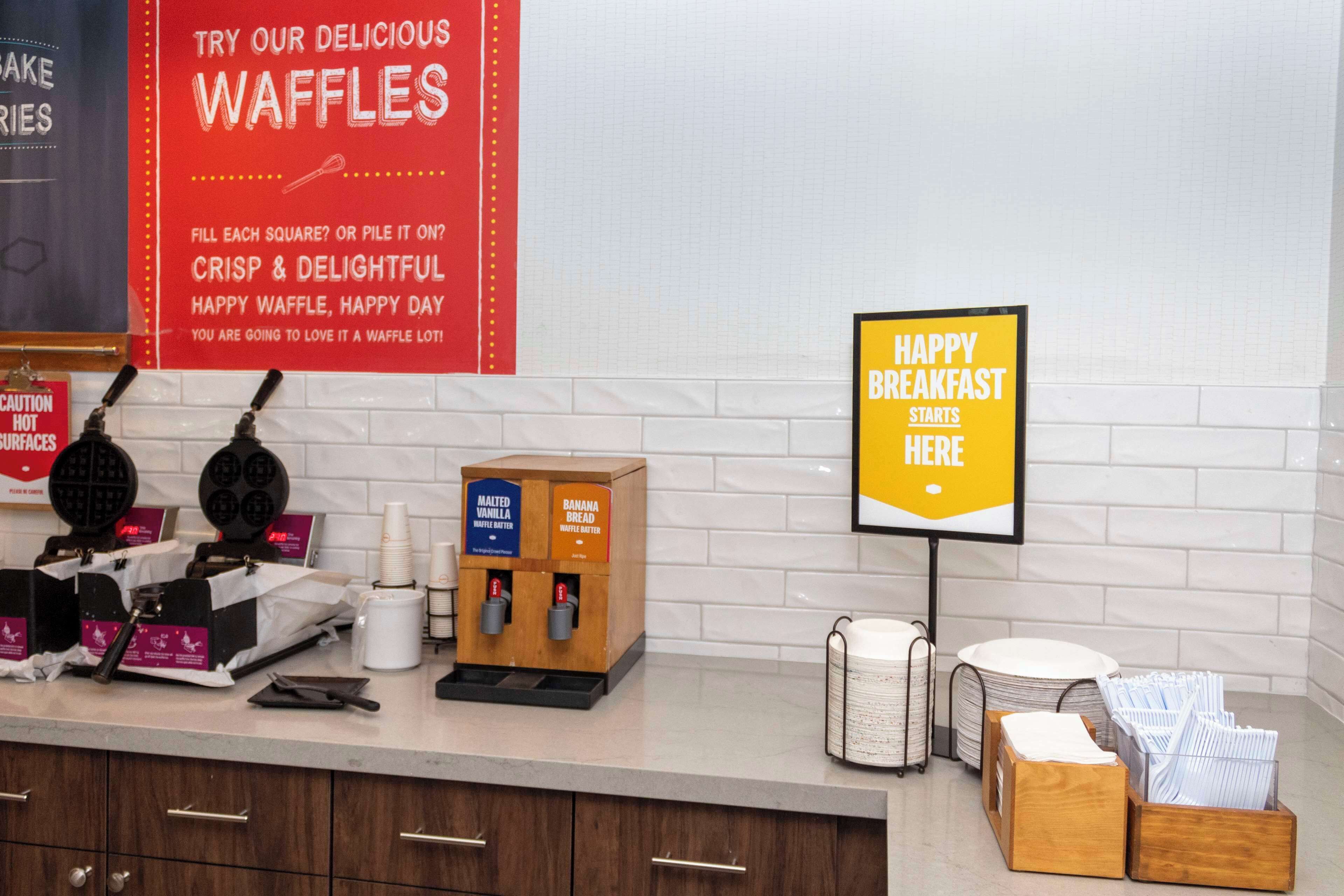 A breakfast area with waffle makers, a sign promoting waffles, and a sign indicating breakfast starts here.