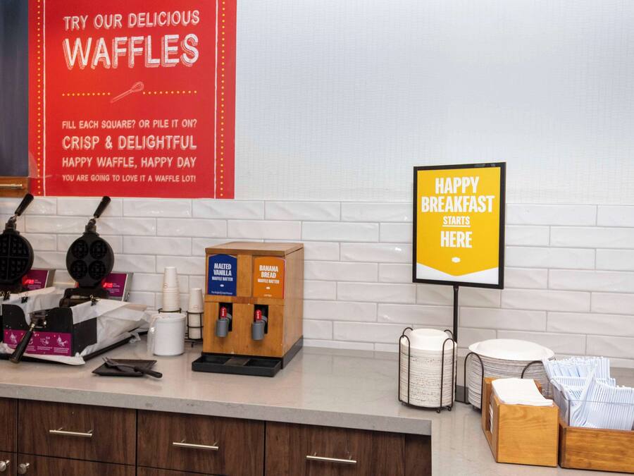 A breakfast area with waffle makers, a sign promoting waffles, and a sign indicating breakfast starts here.