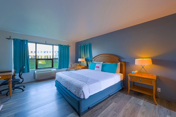 A hotel room with a large bed, a desk, a chair, and a nightstand with a lamp.
