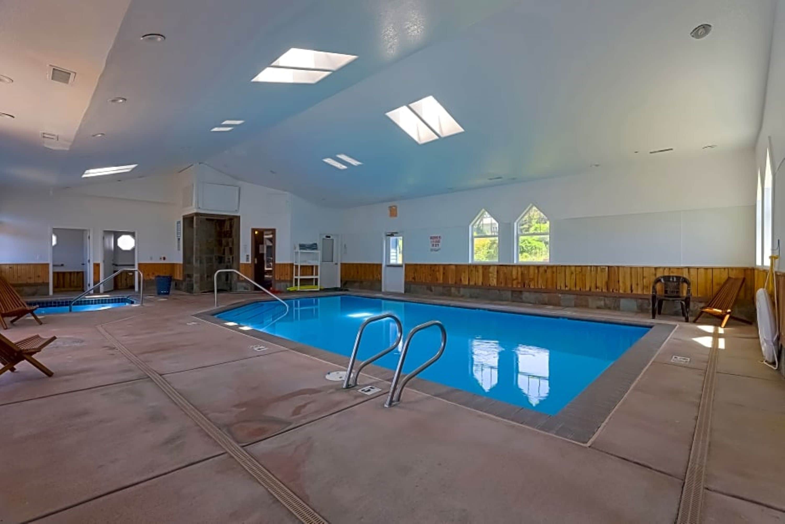 A large indoor swimming pool with clear blue water, surrounded by a concrete deck. There are lounge chairs and a small table near the pool.