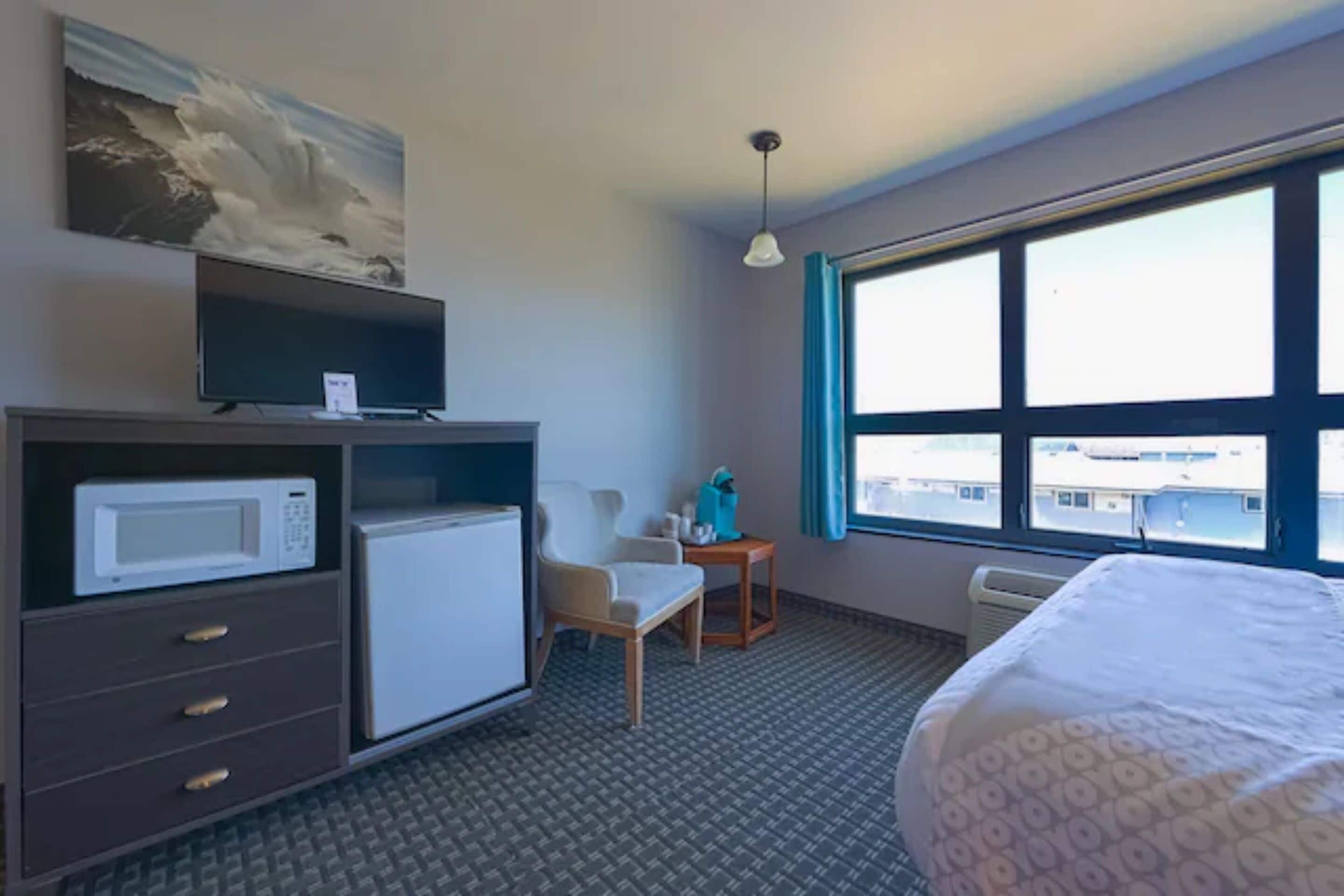 A hotel room with a bed, a desk with a computer, a microwave, a chair, a small table with a lamp, and a large window with a view of buildings.