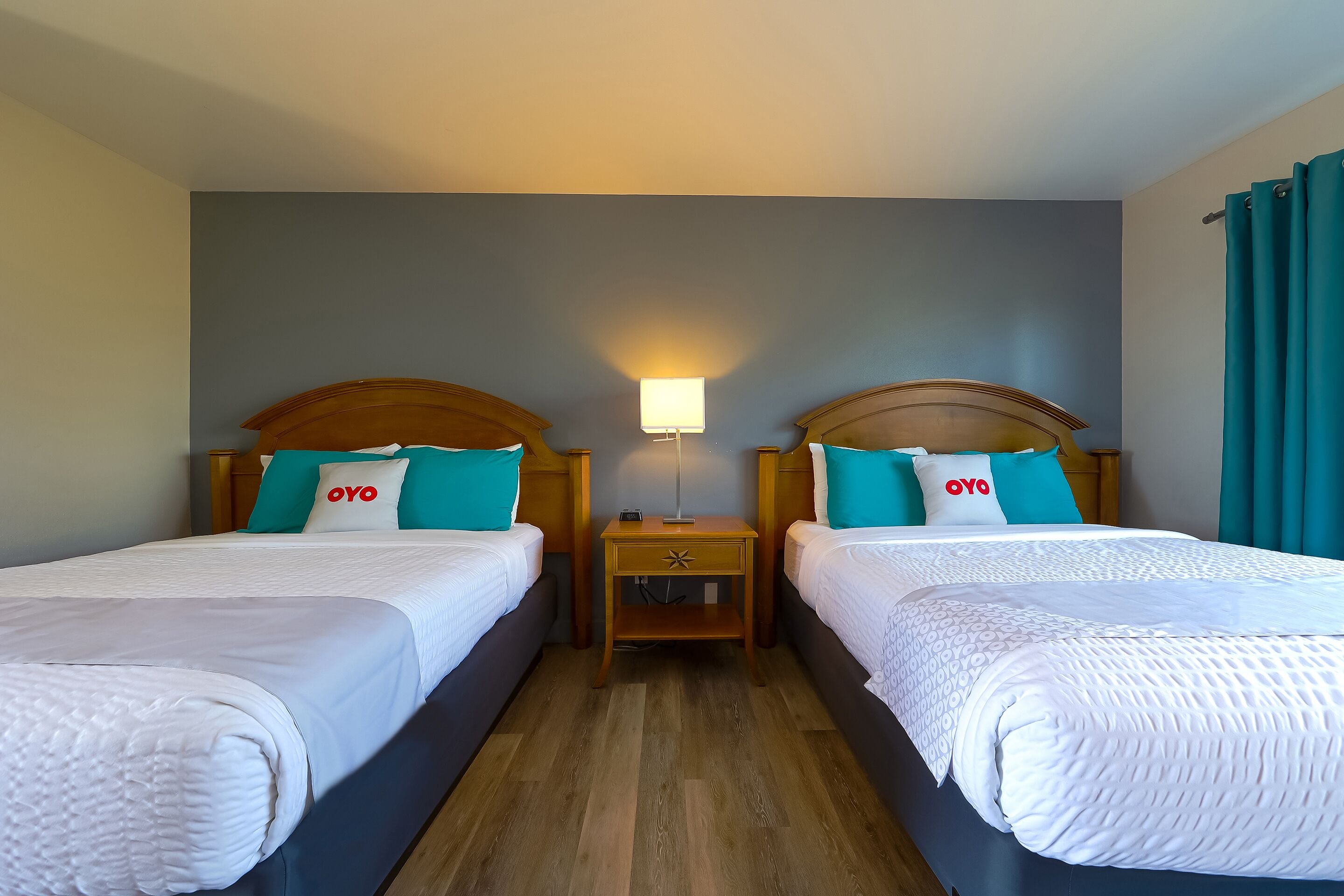 A hotel room with two beds, each with a wooden headboard, white bedding, teal pillows, and a small bedside table with a lamp.
