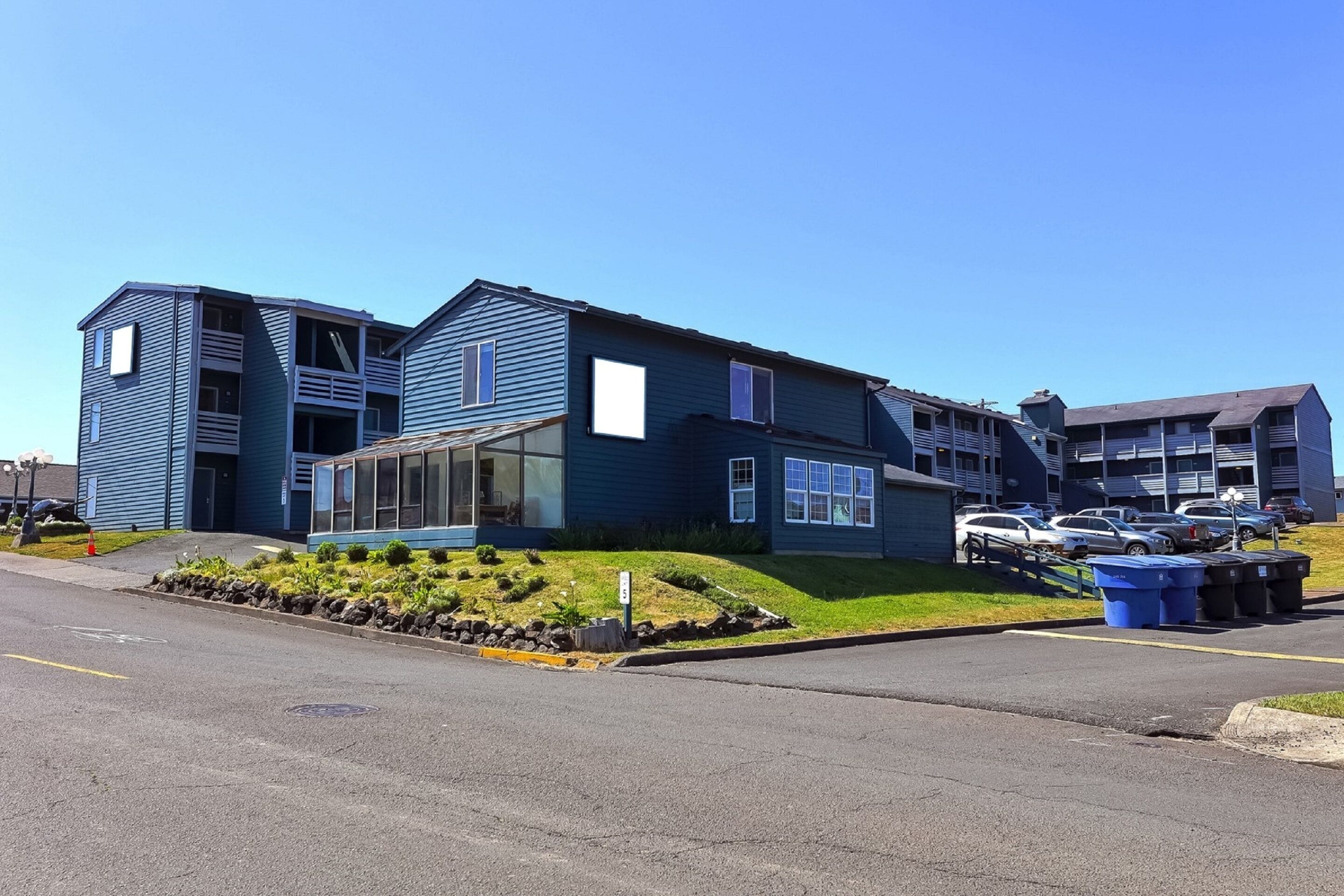 A multi-story residential building with a blue exterior, a landscaped front yard, and a parking area with several cars.