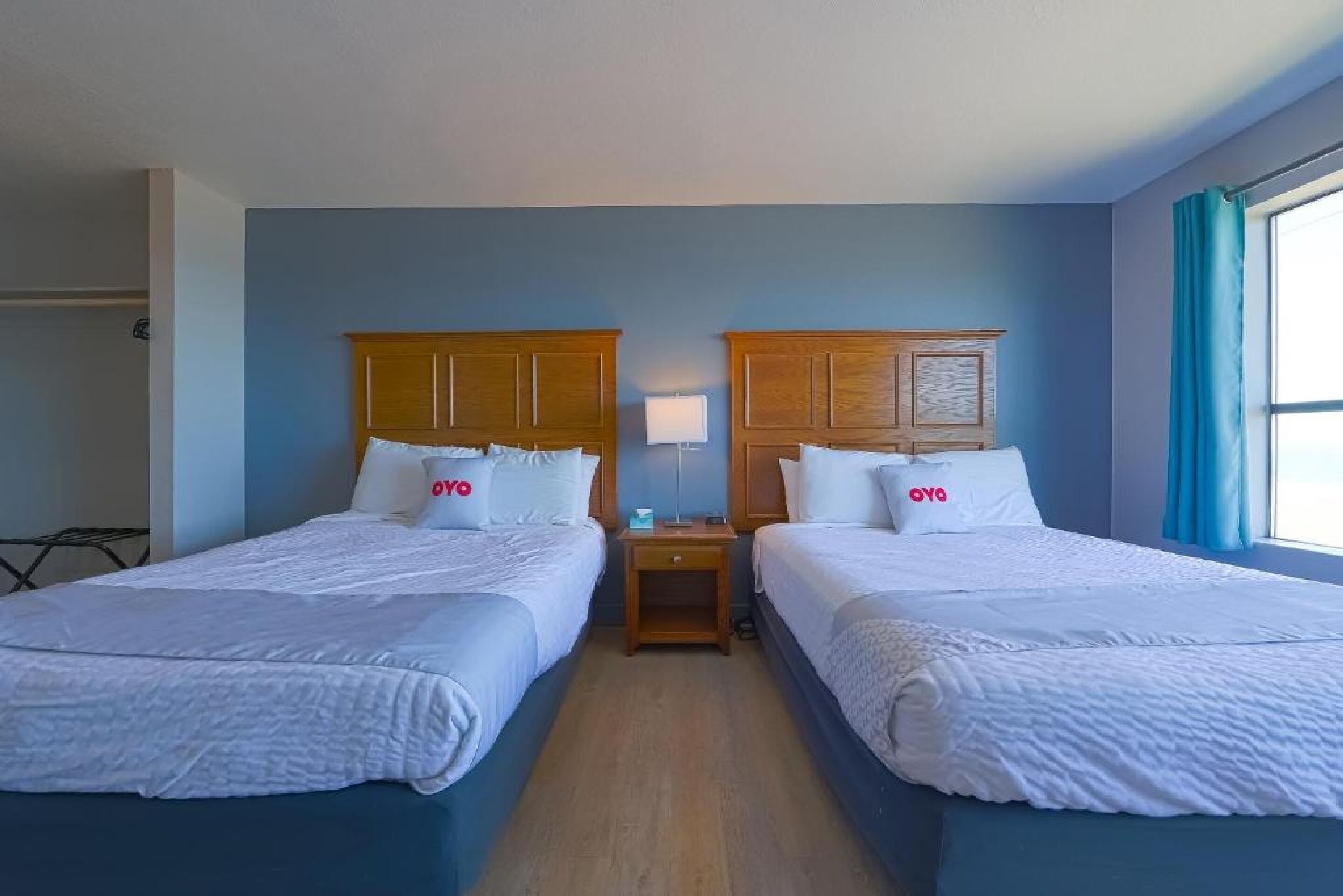 Une chambre d’hôtel avec deux lits, des têtes de lit en bois, du linge de lit blanc, une table de chevet avec une lampe et une fenêtre avec des rideaux.